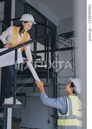 Construction manager and engineer dressed in orange work vests and hard helmets explore construction documentation on the building site near the steel frames 129846002