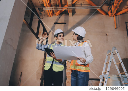 Construction manager and engineer dressed in orange work vests and hard helmets explore construction documentation on the building site near the steel frames 129846005