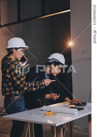 Construction manager and engineer dressed in orange work vests and hard helmets explore construction documentation on the building site near the steel frames Construction manager and engineer dressed in orange work vests and hard helmets explore construction documentation on the building site near the steel frames 129846010
