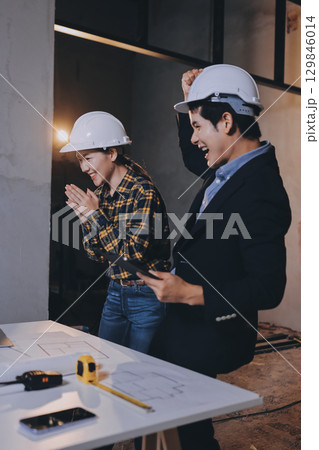 Construction manager and engineer dressed in orange work vests and hard helmets explore construction documentation on the building site near the steel frames Construction manager and engineer dressed in orange work vests and hard helmets explore construction documentation on the building site near the steel frames 129846014