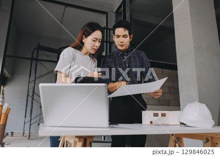 Construction manager and engineer dressed in orange work vests and hard helmets explore construction documentation on the building site near the steel frames 129846025