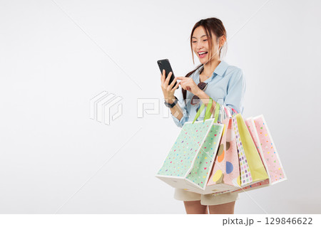 Happy Asian woman using smartphone shopping online holding fashion bags, positive expression on her face in store, holding her new purchase bags, browsing her mobile phone, isolated white background Happy Asian woman using smartphone shopping online holding fashion bags, positive expression on her face in store, holding her new purchase bags, browsing her mobile phone, isolated white background 129846622