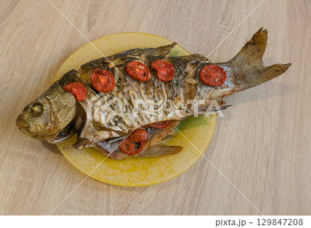 Baked large crucian fish with herbs, tomatoes and spices on a plate on the table. 129847208