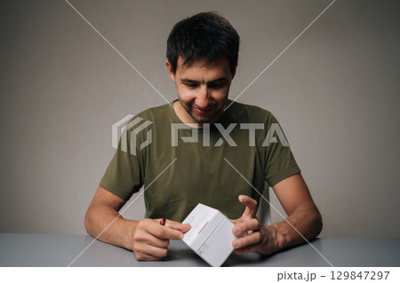 Happy man unpacking new electronic cigarette using small knife sitting at table on isolated background. Concept of modern alternative to traditional smoking. Studio shot, copy space. Happy man unpacking new electronic cigarette using small knife sitting at table on isolated background. Concept of modern alternative to traditional smoking. Studio shot, copy space. 129847297