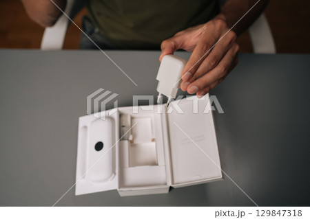 High-angle view of unrecognizable man unpacking box, holding white smartphone charger being unpacked from cardboard box, sitting on gray table. Concept of contemporary gadget unboxing. 129847318