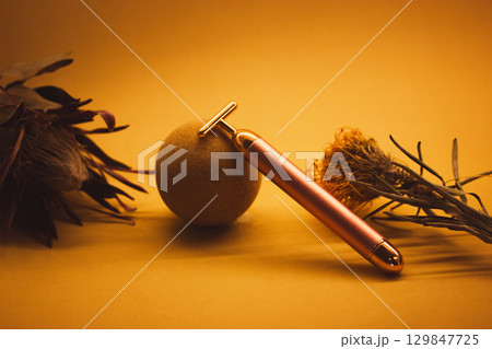 A golden electrical facial T-massager on a yellow background. Beauty care treatment anti-aging product mock-up with dry exotic flowers. Female routine 129847725