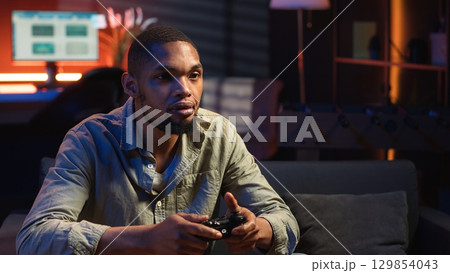 African american man spends free time playing video games with popcorn snack, having fun during game night in his cozy loft. Young adult connecting his console to the television. Camera B. African american man spends free time playing video games with popcorn snack, having fun during game night in his cozy loft. Young adult connecting his console to the television. Camera B. 129854043