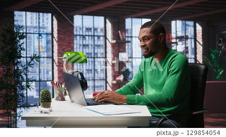 African american male teleworker completing spreadsheets on laptop, entering numerical data into cells and calculating totals. Guy using spreadsheet software functions. Camera B. 129854054