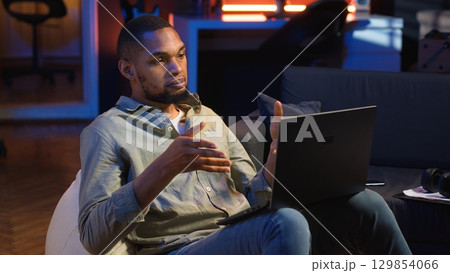 Black man joining online lesson to work on new skills from home space, attend web conference call and watching lecture from loft. Person participates at a virtual training class. Camera B. 129854066