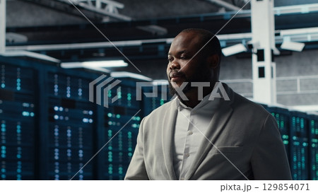 Smiling engineer in server hub monitoring data center energy consumption across components. Happy IT expert walking in data center, making sure rigs sensors are functioning optimally Smiling engineer in server hub monitoring data center energy consumption across components. Happy IT expert walking in data center, making sure rigs sensors are functioning optimally 129854071