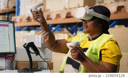 African american woman working with VR tech system to process orders and manage parcel shipment tracking, hands free inventory control. Virtual reality guarantees fast delivery. Camera B. 129854126