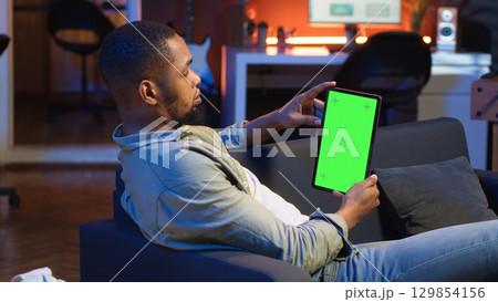 Black remote worker looking at green screen on his device at home, relaxing in the living room and solving freelancing tasks. Guy being casual in trendy apartment, teleworking. Camera B. Black remote worker looking at green screen on his device at home, relaxing in the living room and solving freelancing tasks. Guy being casual in trendy apartment, teleworking. Camera B. 129854156