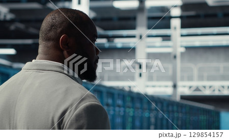 System administrator in data center doing inspection on infrastructure tech handling user requests. African american worker in server farm analyzing traffic patterns of equipment, camera B System administrator in data center doing inspection on infrastructure tech handling user requests. African american worker in server farm analyzing traffic patterns of equipment, camera B 129854157