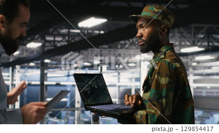 Military technician in data center monitoring warfare drone telemetry on laptop dashboard. IT specialist in army server room using tactical interface on notebook to support aerial operations Military technician in data center monitoring warfare drone telemetry on laptop dashboard. IT specialist in army server room using tactical interface on notebook to support aerial operations 129854417