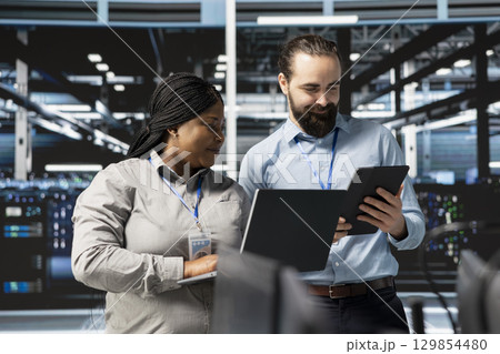 Happy system administrators in data center using devices, verifying firewall configurations. Jolly teamworking IT experts performing equipment vulnerability scans in server hub 129854480