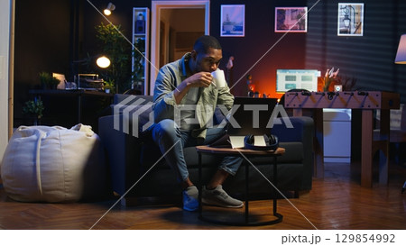 Young black guy working on his freelance tasks in the living room, adjusting his job schedule and enjoying work from his cozy home. Person navigating on web with laptop, multitasking. Camera A. Young black guy working on his freelance tasks in the living room, adjusting his job schedule and enjoying work from his cozy home. Person navigating on web with laptop, multitasking. Camera A. 129854992