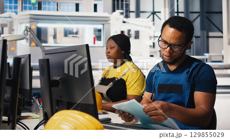 African american engineer manages data from solar machinery on desktop, working in a solar panel manufacturing factory. Male technician in automated renewable energy production line. Camera B. 129855029