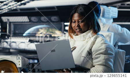 Astronaut returning from planet surface taking helmet off, talking with colleague using laptop to verify parameters. Cosmonaut finishing expedition chatting with coworker updating flight log Astronaut returning from planet surface taking helmet off, talking with colleague using laptop to verify parameters. Cosmonaut finishing expedition chatting with coworker updating flight log 129855141