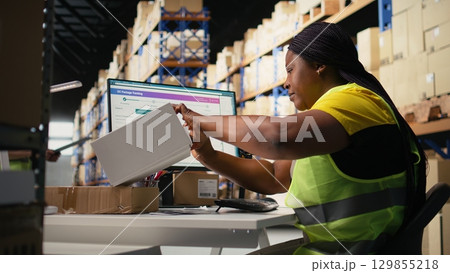 Black woman placing adhesive shipment tags on cardboard boxes, handles labeling and packaging tasks for order processing. Worker applying printed awb labels in storage depot. Camera A. 129855218