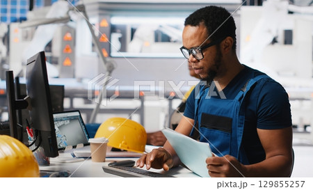 Black engineer implements automation logic in solar factory software, working on manufacturing solar panel designs for alternative energy. High tech production line. Camera A. Black engineer implements automation logic in solar factory software, working on manufacturing solar panel designs for alternative energy. High tech production line. Camera A. 129855257