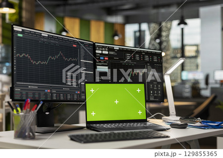 Green screen laptop in brokerage office next to workstation monitor displaying live stock charts. Mockup notebook on desk and computer with financial software in prop firm workspace 129855365