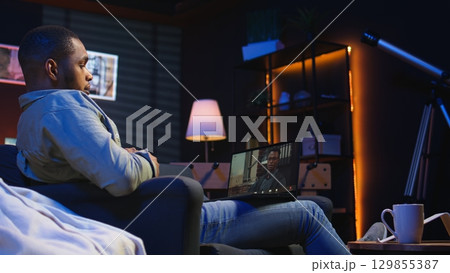Focused black guy smiling and waving during a web conference in his living room, showcasing online discussion in a digital and wireless lifestyle. Conference speaker with attendees on call. Camera A. 129855387