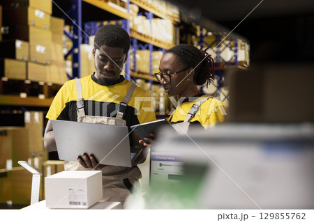 Warehouse workers overseeing parcels tracking info on laptop, preparing pallets for outbound shipment. Organized depot with storage racks, cardboard boxes and inventory processing tools. Warehouse workers overseeing parcels tracking info on laptop, preparing pallets for outbound shipment. Organized depot with storage racks, cardboard boxes and inventory processing tools. 129855762