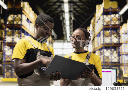Black colleagues preparing orders in boxes for shipment in large warehouse, checking inventory on laptop and managing cargo distribution. Sorting packages for logistics management. 129855823