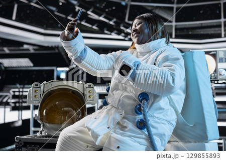 Cheerful female astronaut in starship travelling to space doing selfies using cellphone camera. Happy woman aboard orbit station having fun doing pictures on phone during cosmic journey 129855893