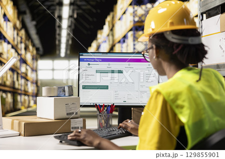 Black woman working in logistics center with tracking info on computer software, updating the route and other shipment details in real time. Cargo parcel status for e-commerce orders. 129855901
