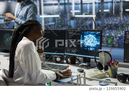 Female programmer in data center answers telephone call, assisting users with AI troubleshooting. Server room IT staff member talks in phone call, managing clients issues using artificial intelligence 129855902