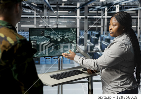 Teamworking army experts in data center confirming target locations using aerial image overlay on monitor. Military units in server room gathering strategic terrain data via satellite systems Teamworking army experts in data center confirming target locations using aerial image overlay on monitor. Military units in server room gathering strategic terrain data via satellite systems 129856019