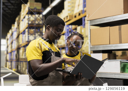 African american team preparing merchandise for dispatch in large warehouse, checking inventory on laptop and managing cargo distribution. Sorting packages for logistics infrastructure. 129856119