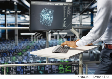 Black software engineer reviews machine learning systems on computer, standing on industrial platform at elevated workstation. Server hub technician monitoring AI infrastructure. Black software engineer reviews machine learning systems on computer, standing on industrial platform at elevated workstation. Server hub technician monitoring AI infrastructure. 129856128