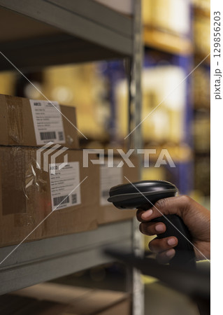 Close up of industrial depot worker scanning parcel details using tracking software, walking near huge storage racks. E-commerce delivery management system ensures accuracy in order processing. 129856203