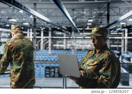Woman in server room command center managing military operations using laptop. African american soldier wearing uniform running diagnostics on infrastructure in army data center 129856207