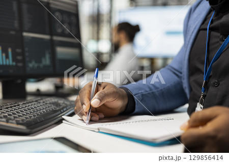 Close up of african american worker writes notes on textbook in the office, analyzing charts and graphs to align company objectives with strategy. Drive progress and measurable performance. Close up of african american worker writes notes on textbook in the office, analyzing charts and graphs to align company objectives with strategy. Drive progress and measurable performance. 129856414