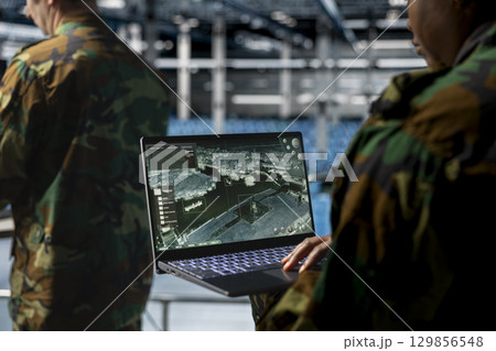 Close up of military technician in data center tracking satellite coordinates using laptop interface. IT specialist in server room analyzing reconnaissance data through satellite imaging systems Close up of military technician in data center tracking satellite coordinates using laptop interface. IT specialist in server room analyzing reconnaissance data through satellite imaging systems 129856548