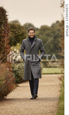 Elegant handsome man walking on formal garden path wears sophisticated long grey coat and turtleneck, radiating sense of serene happiness and confident ease outdoors during autumn 129856875