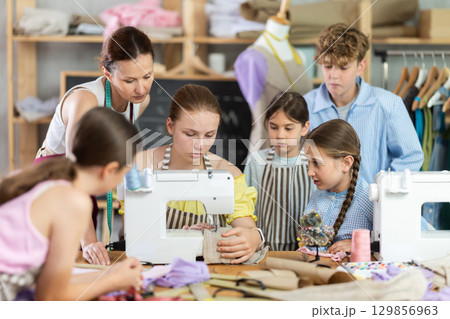 Woman teacher demonstrates how to sew on a sewing machine. Children gather around and watch 129856963