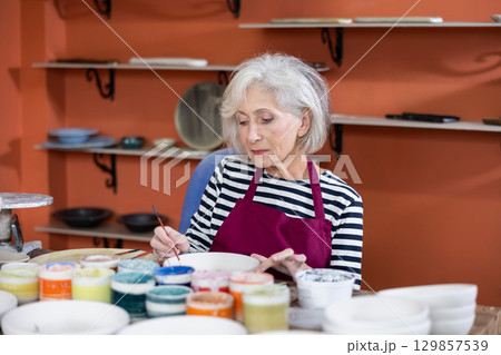 Mature craftswoman painting clay plates acrylic paints in art studio 129857539