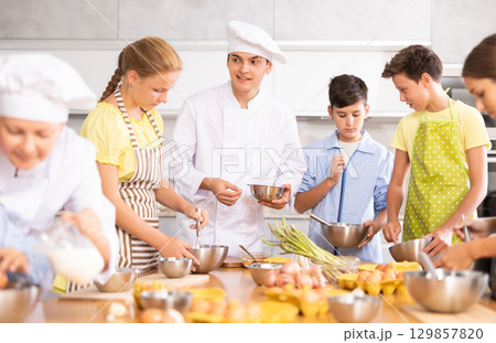 During cooking lesson, male cook tells children about culinary rules, woman assistant helps child 129857820