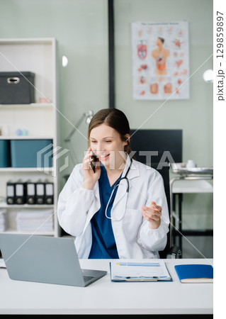Young female doctor in white medical uniform using laptop and tablet talking video conference call at desk,Doctor sitting at desk 129859897
