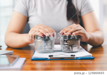 Young Asian woman making stack of coin. invest save finance concept, saving money, investment. dollar bills on table with pile of coins and banks calculator, money to save and invest to make income. 129860685