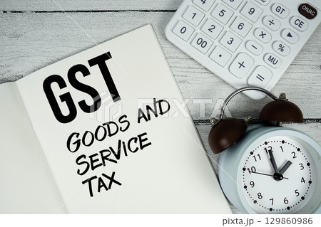 GST Goods and Service Tax text on paper notebook page with alarm clock and calculator on wooden background 129860986