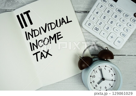 IIT Individual Income Tax text on paper notebook page with alarm clock and calculator on wooden background 129861029