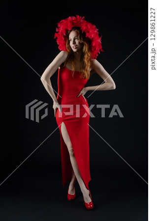 Woman posing in a stunning red dress with a dramatic feathered hat in a studio setting 129861227