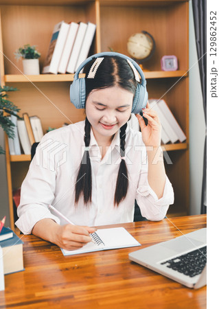 Asian girl student wear headphones learning online watching webinar class looking at laptop computer eLearning remote lesson making notes or video calling virtual conference meeting teacher at home. Asian girl student wear headphones learning online watching webinar class looking at laptop computer eLearning remote lesson making notes or video calling virtual conference meeting teacher at home. 129862452