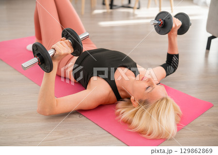 Focused middle aged blonde woman lying on yoga mat performing dumbbell chest press workout for strength and fitness in living room Focused middle aged blonde woman lying on yoga mat performing dumbbell chest press workout for strength and fitness in living room 129862869