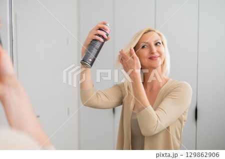 Elegant blonde woman in nude outfit applying hair spray to style and fix her hairstyle while standing by mirror indoors 129862906